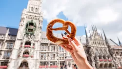 Eine Hand hält eine traditionelle bayerische Breze, im Hintergrund das Münchener Rathaus