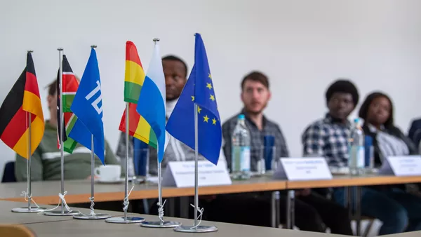 Junge Menschen sitzen an Tischen und hören einem Vortrag zu. Im Vordergrund sind diverse Flaggen zu sehen.
