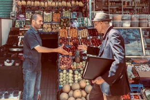 Two men greet each other in front of a fruit stand with a friendly handshake