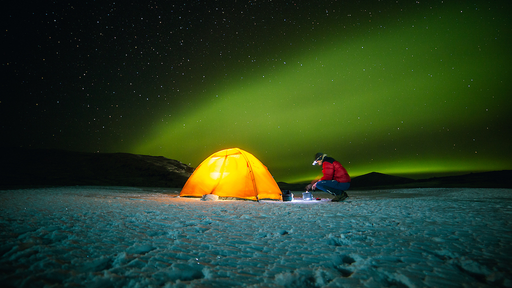 Ein Mann kniet im Dunkeln vor seinem gelb erleuchteten Zelt. Im Hintergrund faszinierender Sternenhimmel und grünes Nordlicht.