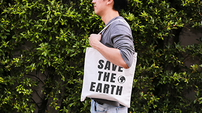 Junger, vor Sträuchern stehender Hipster trägt einen Save The Earth-Jutebeutel über der Schulter